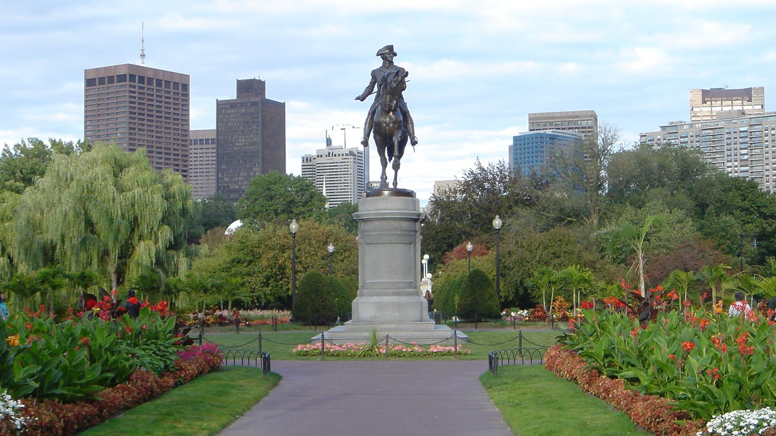A statue of a man on a horse over a public park with green lawns, red flowers, and paved paths with the Boston skyline in the background.