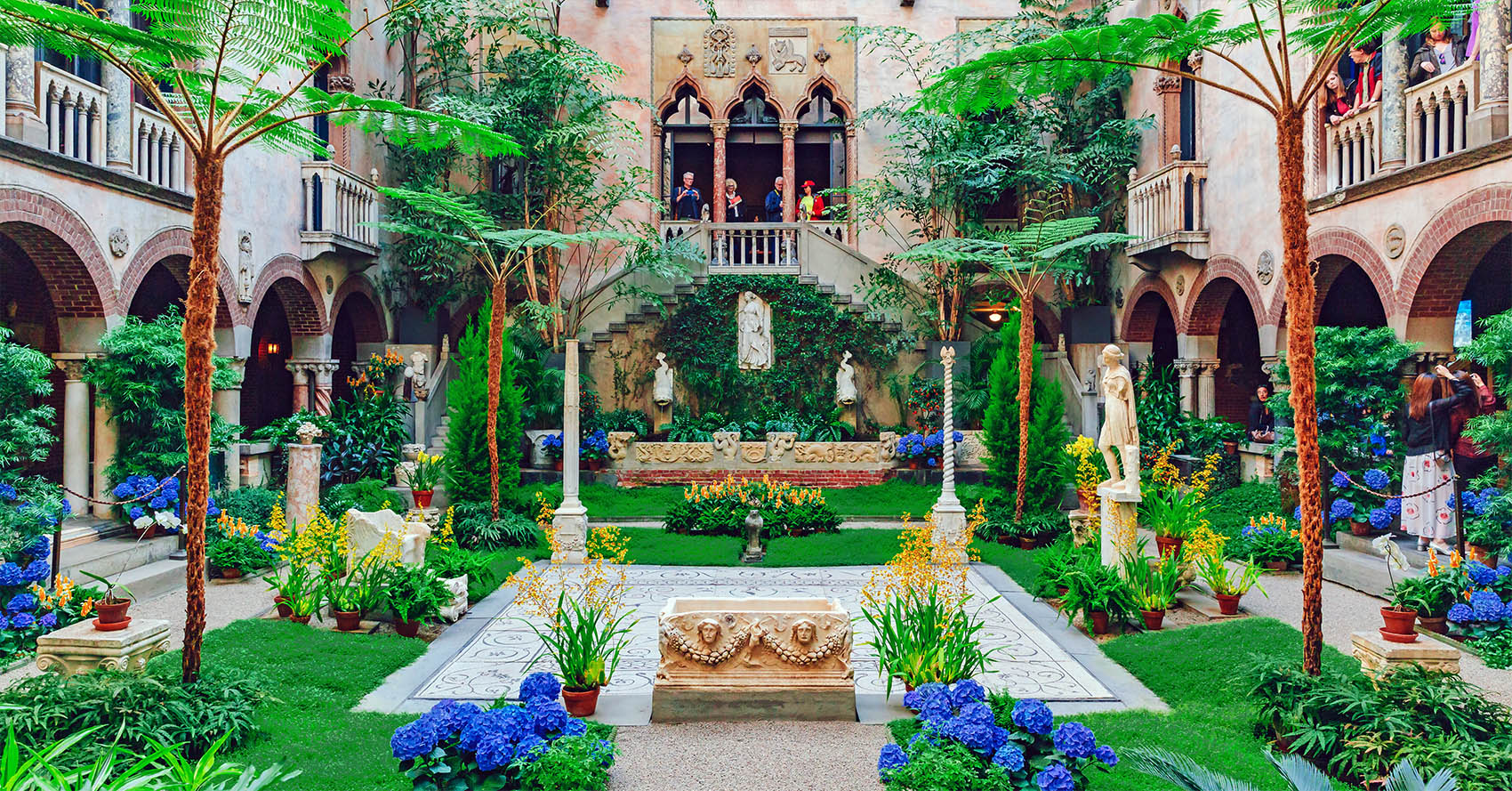A lush green courtyard filled with flowers and with a stone wall and arched windows in the background.